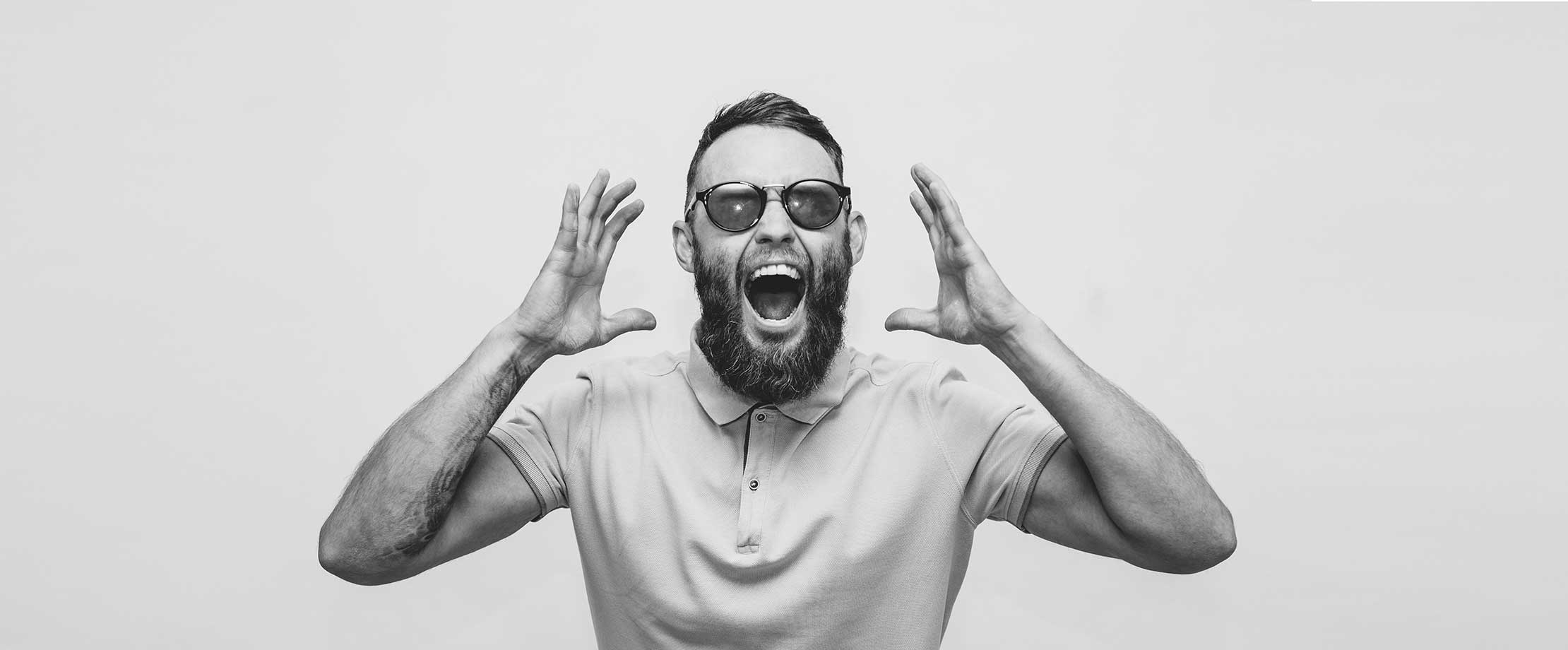 Ein Mann mit Sonnenbrille und Vollbart hat die Hände neben seinem Kopf erhoben und den Mund zum Schrei aufgerissen. Das Bild ist schwarz-weiß.