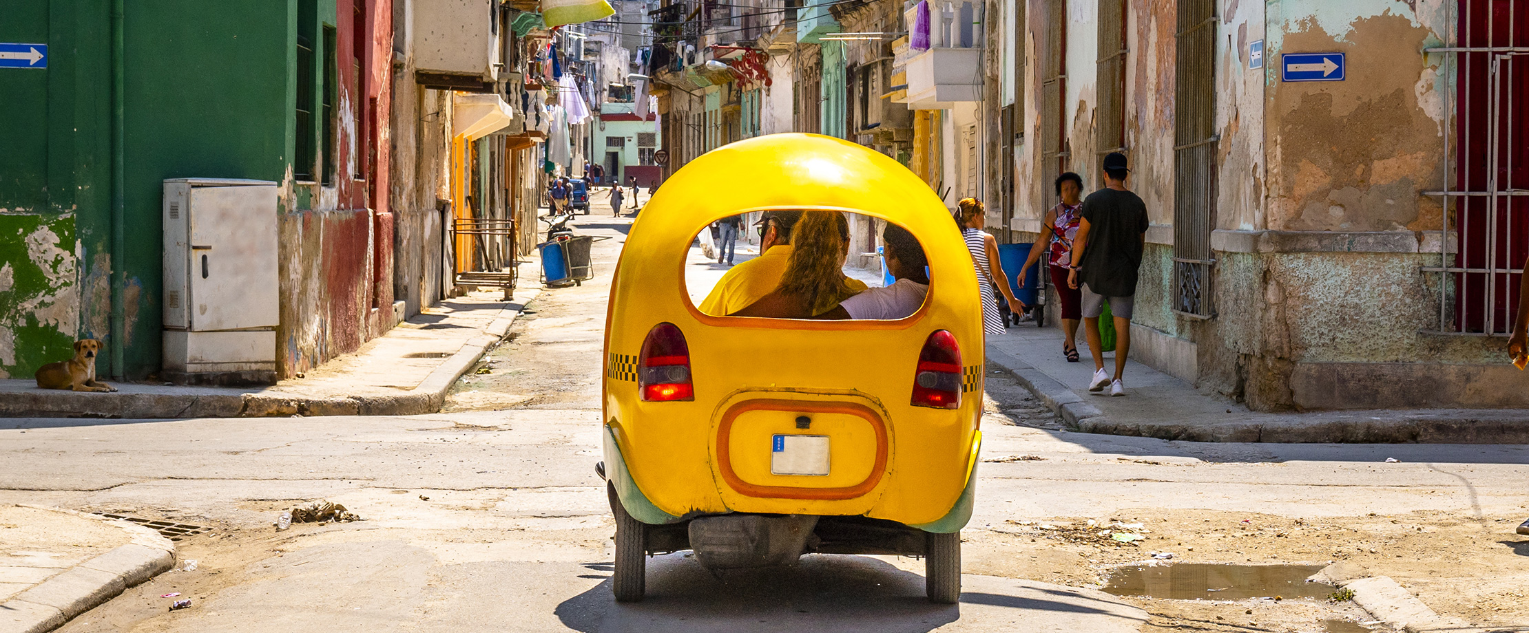 Ein kleines, rundes, gelbes Auto fährt durch eine belebte kubanische Seitengasse. In der Gasse spazieren Menschen in sommerlicher Kleidung.