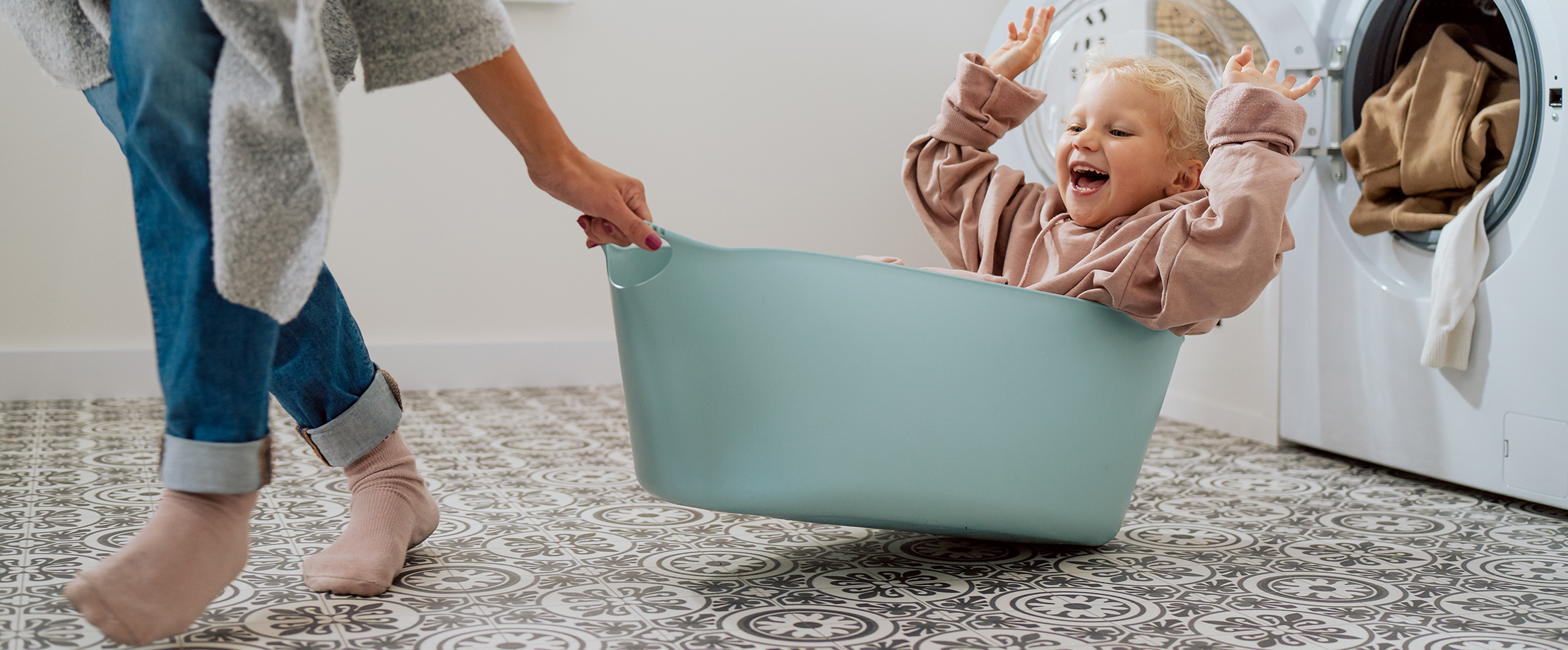 Fröhliches kleines Mädchen sitzt in einem Wäschekorb und wird von der Mutter gezogen